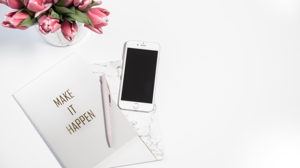A minimalistic desk setup with a planner saying "make it happen," a smartphone, a pen, and a vase of pink tulips on an SQF template background.
